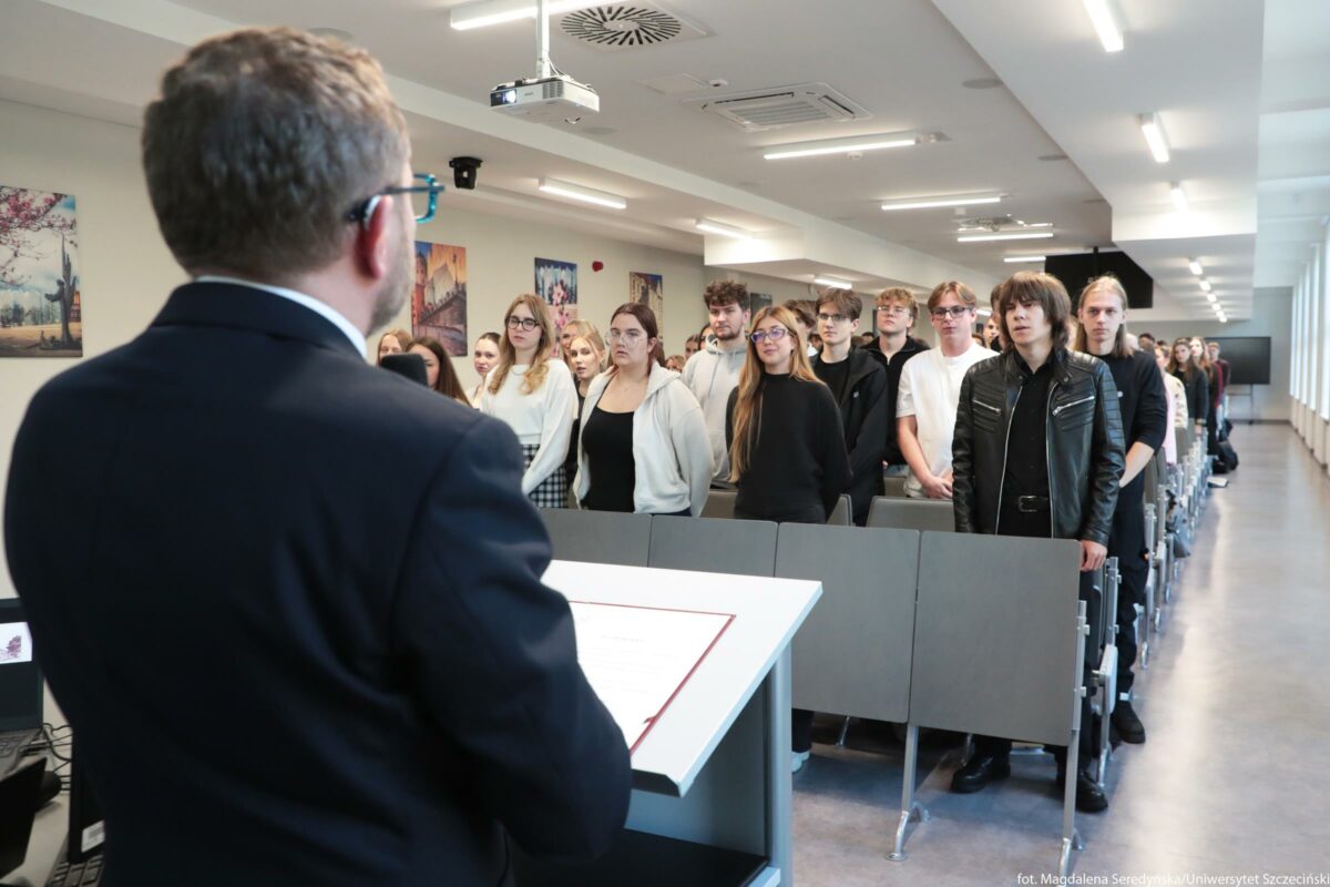 Grupa stojących studentów w sali wykładowej podczas uroczystości. Na pierwszym planie z lewej strony widoczny jest mężczyzna w garniturze, odwrócony tyłem, stojący za jasną mównicą z dokumentem w czerwonej ramce. Przed nim, w kilku rzędach pomiędzy składanymi siedzeniami, stoi kilkadziesiąt młodych osób – kobiety i mężczyźni – większość w casualowych ubraniach: bluzy, swetry, koszule, ciemne spodnie; jedna osoba z przodu ma czarną skórzaną kurtkę, inna jasną bluzę, kilka osób ma okulary. Wszyscy stoją w skupieniu, twarzami zwróceni w stronę mówiącego przy mównicy, tworząc długi rząd aż po tył sali. Pomieszczenie jest nowoczesną, jasną salą uniwersytecką z rzędem okien po prawej stronie, sufitowymi lampami LED i projektorem zawieszonym nad środkiem. Na ścianie po lewej stronie wiszą barwne obrazy przedstawiające miejskie pejzaże.