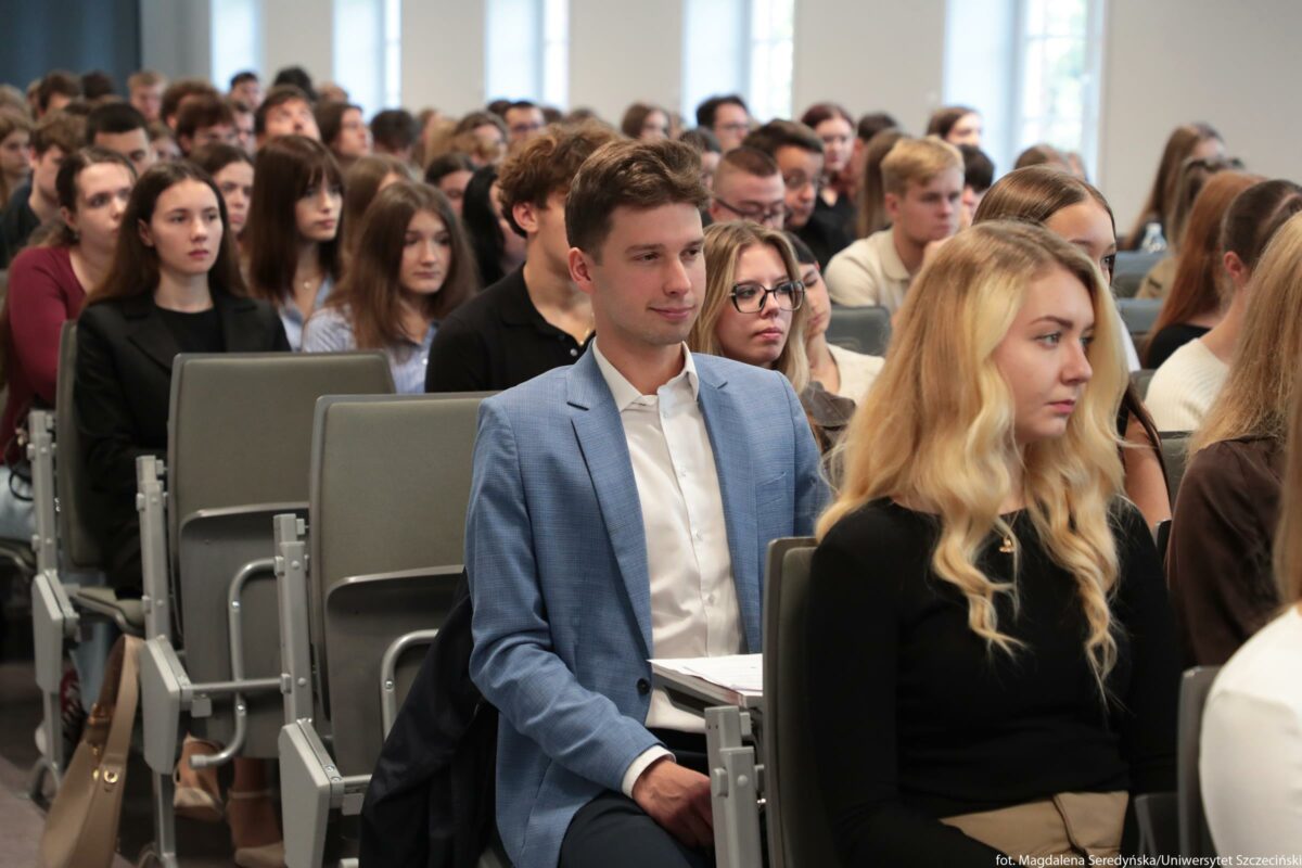 Duża sala wykładowa wypełniona młodymi ludźmi siedzącymi w rzędach foteli. W centrum kadru siedzi młody mężczyzna o jasnej karnacji, w jasnoniebieskiej marynarce i białej koszuli, z krótkimi brązowymi włosami, patrzący przed siebie z lekkim uśmiechem. Na pierwszym planie po prawej stronie znajduje się młoda kobieta o jasnej karnacji i długich, falowanych blond włosach, ubrana w czarną bluzkę; siedzi wyprostowana i patrzy w przód. Za nimi oraz po bokach widać wiele innych osób, głównie młodych kobiet i mężczyzn w casualowych lub półeleganckich ubraniach, wszyscy siedzą w podobnych, składanych fotelach z szarymi oparciami. Twarze osób są skupione i poważne, co sugeruje uczestnictwo w wykładzie lub oficjalnym spotkaniu. Ściany sali są jasne, a w tle widoczne są wysokie prostokątne okna, przez które wpada dzienne światło.