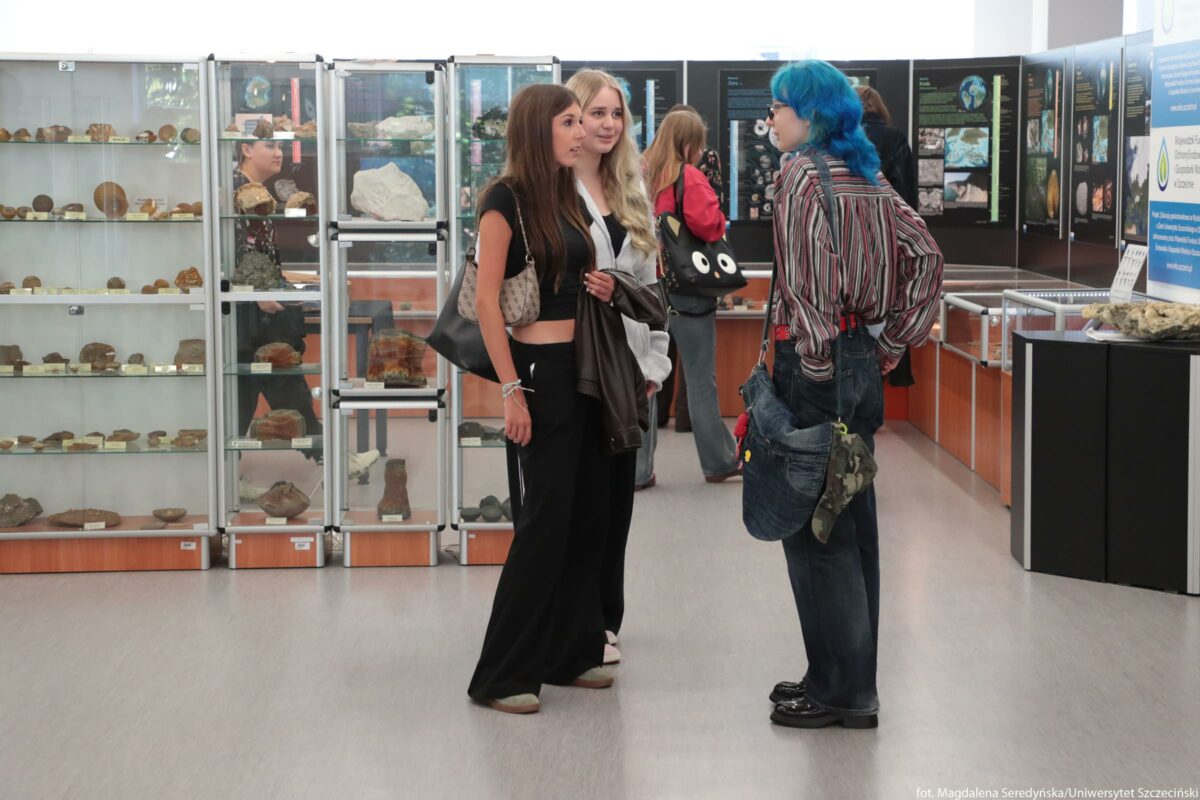 Grupa młodych osób stojąca w muzeum geologicznym. Na pierwszym planie stoją trzy osoby i rozmawiają. Po lewej stoi dziewczyna o długich, prostych, brązowych włosach, w czarnym, krótkim topie i szerokich czarnych spodniach; na ramieniu ma beżową torebkę z jasnym wzorem, w ręku trzyma ciemną kurtkę. Obok niej, lekko z tyłu, stoi dziewczyna o długich, falowanych blond włosach, w czarnym topie i jasnej rozpinanej bluzie, z uśmiechem skierowanym w stronę rozmówcy. Naprzeciwko nich stoi osoba z intensywnie niebieskimi włosami do ramion, w luźnej koszuli w pionowe paski w wielu kolorach i szerokich dżinsach; na ramieniu wisi duża, ciemna torba, do której przypięta jest szmatka w wojskowym, moro wzorze i kilka drobnych przypinek. W tle po lewej stronie widać szklane gabloty wypełnione kamieniami i skamieniałościami ułożonymi na półkach. Po prawej stronie, w głębi, znajdują się tablice wystawowe z kolorowymi grafikami i opisami, a także niskie gabloty z eksponatami geologicznymi. Między gablotami przechodzą jeszcze dwie inne osoby – jedna z czarną torbą z dużymi, białymi oczami jako motywem; obie oglądają wystawę. Podłoga jest jasnoszara, a cała przestrzeń sprawia wrażenie nowoczesnej sali wystawowej.