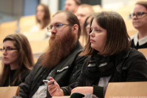 Sala wykładowa. Na pierwszym planie kobieta ze słuchawkami i mężczyzna z długą brodą. W tle kilkoro innych uczestników Dni Adaptacyjnych.