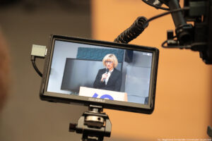 Ujęcie na ekran kamery i rejestrowany przez nią obraz. Na obrazie kobieta z mikrofonem stojąca za mównicą.