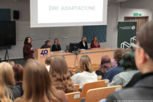 Sala wykładowa (ujęcie bokiem). Na krzesłach w ustawieniu stopniowym studenci – uczestnicy Dni Adaptacyjnych (tyłem do kadru). Na dole po lewej kobieta z mikrofonem przy mównicy z logiem 40-lecia Uniwersytetu Szczecińskiego. Obok, za długim biurkiem, cztery kobiety prowadzące wydarzenie. Po prawej ciemnozielony rollup z logiem Wydziału Nauk Ścisłych i Przyrodniczych. Od góry rozwinięty ekran z napisem Dni Adaptacyjne.