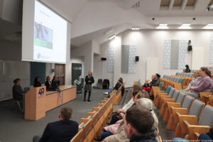 Sala wykładowa (ujęcie bokiem). Po prawej na krzesłach w ustawieniu stopniowym uczestnicy Dni Adaptacyjnych (bokiem do kadru). Po lewej kobieta z mikrofonem przy zerkająca na rozwinięty od góry duży ekran. Obok, za długim biurkiem, cztery kobiety prowadzące wydarzenie. W tle ciemnozielony rollup z logiem Wydziału Nauk Ścisłych i Przyrodniczych.