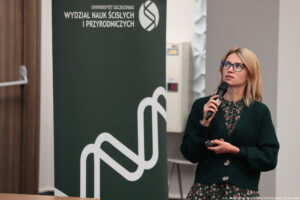 Sala wykładowa. Kobieta z mikrofonem, trzymająca pilot do slajdów. Po lewej ciemnozielony rollup z logiem Wydziału Nauk Ścisłych i Przyrodniczych.