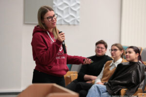 Sala wykładowa. Po lewej studentka w bordowej bluzie z białym logiem Uniwersytetu Szczecińskiego trzyma mikrofon. Po lewej trzy osoby w pierwszym rzędzie podczas Dni Adaptacyjnych.