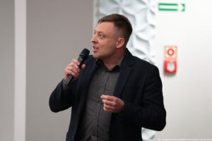 Sala wykładowa. Mężczyzna z mikrofonem (ujęcie bokiem do pasa). Białe tło. Na ścianie znaki ewakuacyjne.