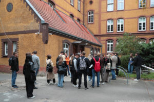 Zabytkowy budynek z cegły, przed którym stoi około 30-osobowa grupa studentów (tyłem do kadru) – uczestników Dni Adaptacyjnych.