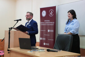 Sala wykładowa (ujęcie bokiem). Po lewej za drewnianą mównicą Prodziekan ds. Studenckich Wydziału Prawa i Administracji – dr hab. Wojciech Staszewski, prof. US. Obok biurku z otwartym laptopem, pilotem do prezentacji i mikrofonem. W tle bordowy rollup z białym logiem WPiA.