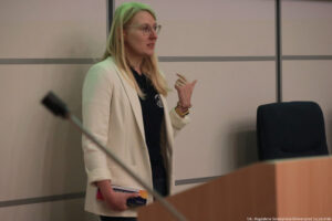 Sala wykładowa. Kierowniczka Akademickiego Biura Karier – Monika Smoguła opowiada uczestnikom Dni Adaptacyjnych o działaniach Biura. Na pierwszym planie (nieostra) mównica z mikrofonem.