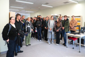 Dwudziestoosobowa grupa studentów odwiedza uczelnianą bibliotekę podczas Dni Adaptacyjnych. W tle półki z książkami.