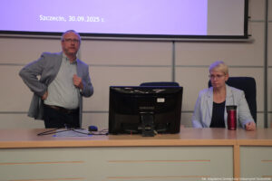 Sala wykładowa. Prelekcja. Na środku długie biurko. Na nim czarny monitor. Po lewej stojący mężczyzna, po prawej siedząca kobieta patrząca na monitor. W tle duży ekran z prezentacją.