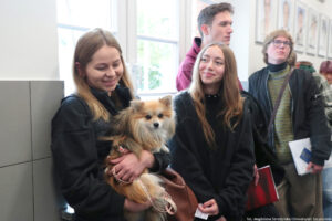 Korytarz uczelni. Czworo studentów. Na pierwszym planie dziewczyna trzymająca pieska i uśmiechająca się do niego. W tle okno i portrety.