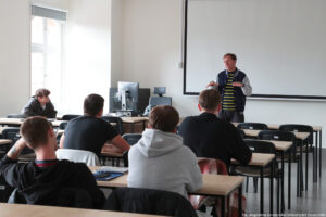 Sala wykładowa. Pięcioro studentów siedzi w ławkach (tyłem do kadru). Przodem wykładowca. Za nim duży biały ekran. Po lewej okno oraz biurko z monitorem i komputerem.