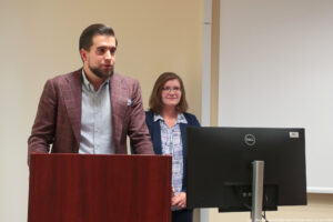 Sala wykładowa. Po lewej za mównicą wykładowca. Za nim kobieta – prodziekan do spraw studenckich. W tle duży biały ekran.