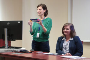 Sala wykładowa. Po lewej czarny monitor. Obok za biurkiem stoi przedstawicielka Akademickiego Biura Karier. Dalej (po prawej) siedzi prodziekan do spraw studenckich Wydziału Humanistycznego. Na biurku mikrofony, pilot, kartki.