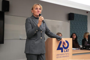 Sala wykładowa. Kobieta trzymająca mikrofon i oparta o drewnianą mównicę z granatowym logiem 40-lecia Uniwersytetu Szczecińskiego. W tle za długim biurkiem trzy inne prowadzące Dni Adaptacyjne dla studentów.