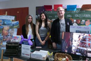 Trzy młode osoby stoją za stołem na szkolnym lub uczelnianym stoisku informacyjnym, na tle tablicy i drzwi. Od lewej stoi młoda kobieta z długimi, prostymi jasnobrązowymi włosami, w białej bluzce i czarnym kardiganie. W środku stoi młoda kobieta o długich czarnych włosach, w szarym topie na ramiączkach i jeansach; ma widoczne tatuaże na przedramionach. Po prawej stoi młody mężczyzna o krótkich jasnych włosach i okularach, w czarnej bluzie/kurtce i szarej koszulce z nadrukiem. Na stole leży czarna maszyna do pisania, stojak z kartką „Uniwersytet Szczeciński / Koło Naukowe / Mediów i Cywilizacji / Medium Kulturalne / Wydział Humanistyczny / Dni otwarte”, fioletowy balon w kształcie pieska, butelka wody, pudełko z napisem „PRAWDA CZY FAKE NEWS”, kubek z długopisami oraz koszyk ze słodyczami. Po prawej stronie stoi monitor pokazujący ulicę w mieście. Po lewej na tablicy/korku wiszą zdjęcia krajobrazów i kartka z napisem „… CZY AI?” (pierwsze słowo jest ucięte). Z tyłu na tablicy jest oś czasu z datami „1450”, „1876”, „1926”, „1969”, „2004” oraz kolorowe kartki z napisem „MEDIUM IS THE MESSAGE” i „MEDIUM IS THE BEST PRZEKAZ…”. Na monitorze/przy nim jest kartka „NASZE WYJAZDY I ZAJĘCIA”. W prawym dolnym rogu widoczny jest napis „fot. Magdalena Seredyńska / Uniwersytet Szczeciński”.