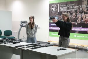 Sala szkoleniowa z dwoma młodymi kobietami i stołami, na których leży wiele sztuk broni palnej. Po prawej stronie stoi kobieta o jasnej cerze, długich jasnobrązowych włosach i okularach. Ma na sobie czarny sweter i szare jeansy. Trzyma pistolet oburącz, z wyprostowanymi rękami, celując w lewo. Po lewej stronie stoi druga kobieta o jasnej cerze, długich brązowych włosach i okularach. Ma na sobie szarą bluzę z kapturem i jasne jeansy. Stoi z rękami z tyłu, patrząc w stronę osoby celującej. Na pierwszym planie są dwa stoły z rozłożoną bronią: kilka długich karabinów/strzelb (z widocznymi lufami i kolbami) oraz co najmniej jeden pistolet leżący po prawej stronie na blacie. W tle widać bieżnię treningową oraz duży baner z napisem: „UNIWERSYTET SZCZECIŃSKI” oraz „INSTYTUT NAUK O POLITYCE I BEZPIECZEŃSTWIE”. Na dole po prawej jest mały podpis: „fot. Magdalena Seredyńska/Uniwersytet Szczeciński”.