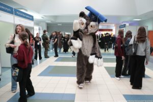 Duży kostium pluszowego zwierzęcia (przebranie z człowiekiem w środku), stojący na środku hali i trzymający na głowie niebieski biret z napisem „Uniwersytet Szczeciński”. Postać ma duże, okrągłe oczy i białe „łapy”, a na nogach fioletowe trampki. W tle widać wnętrze budynku (hol) z grupami ludzi stojących i rozmawiających; część osób jest ubrana na czarno jak na uroczystość akademicką. Po lewej stronie stoją stoiska informacyjne z tablicami; na jednej z nich można odczytać „Wydział Nauk Społecznych”. Na dole zdjęcia widać podpis: „fot. Magdalena Seredyńska/Uniwersytet Szczeciński”.