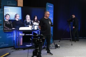Studio nagraniowe z dużym ekranem telewizyjnym i sprzętem kamerowym. Na pierwszym planie stoi mężczyzna w okularach, w czarnej koszulce z logo „UNIWIZJA”, bokiem do kadru. Po prawej stronie inny mężczyzna obsługuje kamerę na statywie. Na dużym ekranie po lewej widać cztery młode kobiety siedzące na kanapie; wyglądają na uczestniczki nagrania lub rozmowy. Tło jest w odcieniach granatu i czerni, z powtarzającymi się napisami „UNIWIZJA” oraz adresem „UNIWIZJA.USZ.EDU.PL”. W dolnej części obrazu widać dodatkową kamerę na statywie, kable i elementy oświetlenia. Tekst w prawym dolnym rogu: „fot. Magdalena Seredyńska / Uniwersytet Szczeciński”.