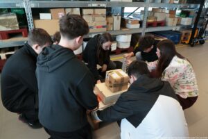 Grupa sześciu młodych osób klęczących i pochylonych nad kilkoma kartonowymi paczkami na podłodze w magazynie. Większość ma ciemne włosy i ubrania w ciemnych kolorach; jedna osoba ma jasną bluzę z kwiatowym wzorem, a jedna osoba ma okrągłe okulary. Na paczkach widać taśmę pakową i etykiety, a jedna osoba trzyma rolkę taśmy i wygląda na to, że okleja lub zabezpiecza paczkę. W tle są metalowe regały z wieloma kartonami i pojemnikami oraz żółte linie na podłodze. W prawym dolnym rogu widoczny jest napis: „Fot. Magdalena Seredyńska/Uniwersytet Szczeciński”.