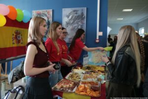 Stoisko z przekąskami na uczelnianym korytarzu, obok którego stoją młode kobiety, a w tle wisi flaga Hiszpanii i kolorowe balony. Na pierwszym planie po lewej stoi młoda kobieta o jasnej skórze i blond włosach do ramion, ubrana w bordową bluzkę bez rękawów i ciemną spódnicę. Obok niej stoją dwie młode kobiety w czerwonych koszulkach; jedna ma jasne włosy, druga ciemne włosy i okulary. Po prawej stronie, przy stole, widać kobietę o jasnej skórze z bardzo długimi blond włosami w czarnej skórzanej kurtce; obok niej częściowo widać osobę z długim warkoczem. Na stole leżą tacki i pudełka z różnymi przekąskami: chipsy, słone paluszki, małe kanapki lub koreczki, ciastka i inne drobne jedzenie, a także miseczki z sosami. Jedna z kobiet w czerwonej koszulce podaje żółty talerzyk w stronę osoby po prawej. W tle na ścianie są plakaty, a przy stole stoi tablica lub stojak z mapą (prawdopodobnie mapą Polski). W prawym dolnym rogu jest mały podpis fotograficzny, ale nie jest w pełni czytelny.