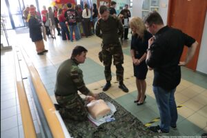 Scena szkoleniowa z pierwszej pomocy na korytarzu budynku. Na podłodze na wojskowym kocu leży fantom tułowia do resuscytacji, a obok klęczy mężczyzna w mundurze moro i wykonuje na nim czynności ratownicze. Obok stoją trzy osoby obserwujące: drugi mężczyzna w moro, kobieta w ciemnej sukience i mężczyzna w czarnej koszuli oraz okularach. W tle widać grupę ludzi zgromadzonych przy stanowisku z żółto-czerwonymi balonami; po lewej są drzwi wejściowe ze szkła. Na dole po prawej jest podpis na zdjęciu: „fot. Magdalena Seredyńska/Uniwersytet Szczeciński”.