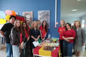 Grupa osób stojących przy stoisku z jedzeniem w budynku, przy niebieskiej ścianie. W kadrze jest około dziesięciu osób, głównie młode kobiety i dwie starsze kobiety; większość ma jasną karnację. Kilka osób ma na sobie czerwone koszulki z żółtym nadrukiem, inne mają ciemne ubrania; jedna osoba z przodu ma czarną skórzaną kurtkę. Na stole leży czerwono-żółta flaga, a na niej tace i pojemniki z przekąskami, talerzyki i drobne akcesoria. Z lewej strony widać kolorowe balony (różowe, pomarańczowe, żółte) oraz fragment flagi Hiszpanii w tle. Na ścianie wiszą dwa plakaty z portretami. W prawym dolnym rogu widoczny napis: „fot. Magdalena Seredyńska/Uniwersytet Szczeciński”.