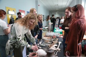 Grupa ludzi przy stołach wystawowych w uczelnianym korytarzu. Na pierwszym planie stoi długi stół przykryty ciemnym obrusem, na którym leżą różne przedmioty: kości (w tym duże, długie kości), fragmenty czaszki lub model czaszki, drobne kości i elementy przypominające zęby, kamienne narzędzia lub odłupki, kawałki ceramiki, sznur lub pleciony materiał, oraz plastikowe pudełka z przegródkami na drobne eksponaty. Po lewej stronie kobieta z kręconymi jasnymi włosami pochyla się nad stołem i układa lub ogląda małe elementy. Po prawej stronie dwie młode kobiety z rudymi włosami stoją obok stołu i rozmawiają, patrząc na eksponaty. W tle widać więcej osób, kolejne stoliki z materiałami informacyjnymi oraz plakaty na ścianach. Na dole zdjęcia widoczny jest podpis: „fot. Magdalena Seredyńska/Uniwersytet Szczeciński”.