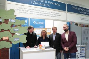 Stoisko targowe uczelni z czterema osobami stojącymi za ladą, na tle banerów „Uniwersytet Szczeciński” oraz napisu „Wydział Ekonomii, Finansów i Zarządzania”. W kadrze widać cztery osoby: trzy osoby w marynarkach po prawej stronie i jedną osobę w ciemnej koszuli po lewej. Na ladzie stoi otwarty laptop oraz kilka słoików z żółtą zawartością. Po lewej stronie zdjęcia znajduje się dekoracja w formie dużego drzewa z zielonymi „liśćmi” i przyczepionymi prostokątnymi kartkami przypominającymi banknoty. W górnej części są listy kierunków/specjalności (m.in. „ekonomia”, „ekonomiczno-prawny”, „finanse i rachunkowość”, „Informatyka i ekonometria”, „gospodarka nieruchomościami”, oraz po angielsku „Economics and IT Applications”, „Business Intelligence w gospodarce”). W prawym dolnym rogu jest podpis fotograficzny: „fot. Magdalena Seredyńska / Uniwersytet Szczeciński”.