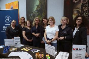 Grupa ośmiu kobiet stojących za stołem promocyjnym na tle ściany z dużymi obrazami. Po lewej stronie jest niebieski roll-up z logo i napisem „UNIWERSYTET SZCZECIŃSKI” oraz „DZIAŁ …” (dalszy tekst częściowo zasłonięty). Kobiety stoją w jednym rzędzie, większość w ciemnych ubraniach; dwie mają białe koszulki lub białą bluzkę. Kilka osób ma okulary; jedna osoba ma okulary przeciwsłoneczne na głowie. Na stole leżą materiały informacyjne i gadżety: ulotki, broszury, naklejki, zakładki, smycze, miski z cukierkami oraz kilka stojaków z kartkami. Widoczne napisy na stojakach obejmują: „STYPENDIA DLA STUDENTÓW”, „STYPENDIUM JUŻ NA I ROKU? TO MOŻLIWE! POROZMAWIAJMY”, „informator”, „MOST PROGRAM MOBILNOŚCI STUDENTÓW I DOKTORANTÓW” oraz „STUDIUJ GDZIE CHCESZ!”. W lewym dolnym rogu zdjęcia jest podpis: „fot. Magdalena Seredyńska/Uniwersytet Szczeciński”.