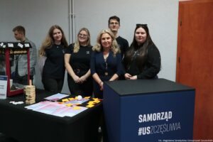 Grupa pięciu osób stojących za stołem na wydarzeniu wewnątrz budynku. Trzy kobiety i jeden mężczyzna mają na sobie czarne koszulki z małym białym logo, a pośrodku stoi kobieta w granatowej sukience; wszyscy patrzą w stronę aparatu. Po lewej stronie w tle widać jeszcze jedną osobę przy czerwonej maszynie (wygląda jak urządzenie do popcornu). Na stole leżą ulotki i kartki, pomarańczowe karteczki samoprzylepne, biała piłeczka oraz drewniana wieża z klocków. Z przodu po prawej stoi duża niebieska skrzynia z napisem „SAMORZĄD #USZCZĘŚLIWIA”. W prawym dolnym rogu jest podpis: „fot. Magdalena Seredyńska/Uniwersytet Szczeciński”.