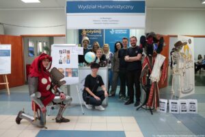 Stoisko informacyjne „Wydział Humanistyczny” z napisem u góry oraz grupą osób pozujących do zdjęcia w holu budynku. Na górnym panelu widoczny jest tekst: „Wydział Humanistyczny”, a pod nim lista kierunków: „archeologia”, „historia”, „media i cywilizacja”, „stosunki międzynarodowe”. W tle widać baner „Uniwersytet Szczeciński”. Przed stoiskiem stoi kilka osób w codziennych ubraniach; jedna osoba kuca z przodu. Po lewej stronie znajduje się osoba w stylizowanym stroju rycerskim: metalowe elementy zbroi, czerwony kaptur i czerwony materiał z dużymi białymi kropkami. Po prawej stronie stoi manekin ubrany w bogato zdobiony, czerwono-złoty strój, obok widoczny jest szkielet anatomiczny i tablica z rysunkiem postaci. Na ladzie stoi globus. Przy podłodze po prawej leżą tabliczki z kodami QR oraz podpisami, m.in. „ARCHEOLOGIA” i „HISTORIA”. W prawym dolnym rogu zdjęcia widnieje podpis: „fot. Magdalena Seredyńska / Uniwersytet Szczeciński”.