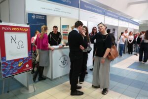 Stoisko informacyjne na dniu otwartym uczelni, z kilkoma osobami rozmawiającymi przy ladzie. W tle widoczne są modułowe ścianki z banerami „Uniwersytet Szczeciński” oraz nazwy wydziałów, m.in. „Wydział Humanistyczny” i „Wydział Nauk Społecznych”. Na ladzie stoi plakat „Teologia” z kolorowymi hasłami. Po lewej stronie znajduje się stojąca tablica z dużym napisem „PŁYŃ POD PRĄD!” i prostym rysunkiem na białym polu. W pierwszym planie stoi kilka młodych osób w ubraniach codziennych; jedna osoba w czarnym stroju rozmawia z osobą o kręconych włosach, a obok stoi osoba w czarnym T-shircie i długiej jasnej spódnicy z brązową torbą na ramieniu. Na dole po prawej widać napis kredytowy: „fot. Magdalena Seredyńska/Uniwersytet Szczeciński”.