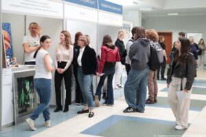 Stoisko informacyjne na uczelnianym korytarzu, przy którym stoi grupa ludzi podczas wydarzenia. W tle widać kilka stanowisk z białymi panelami i niebieskimi nagłówkami; na jednym z nich jest czytelny napis „Wydział Nauk Społecznych”. Po lewej stronie stoi lada z plakatem przedstawiającym twarz osoby w zielonkawym świetle. Przy stoisku rozmawiają osoby w różnym wieku, ubrane na co dzień (jeansy, bluzy, kurtki); część stoi w kolejce lub w skupiskach. Na pierwszym planie po prawej stronie stoi młoda kobieta w czarnej kurtce i jasnych spodniach, trzymająca kartkę. Na dole po prawej jest mały napis ze źródłem zdjęcia: „fot. Magdalena Seredyńska/Uniwersytet Szczeciński”.