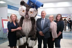 Grupa osób pozująca do zdjęcia obok dużej maskotki-sowy w birecie. Maskotka stoi na środku; ma brązowo-białe futro, duże pomarańczowe oczy i niebieski biret z chwostem oraz napisem „US SZCZECIŃSKI”. Po lewej stronie stoi jasnowłosa kobieta w ciemnej sukience, trzyma dłoń na ramieniu maskotki. Po prawej stronie stoją trzy osoby: siwowłosy mężczyzna w garniturze oraz dwie kobiety (jedna w jasnej marynarce i okularach, druga w ciemnej marynarce i okularach). Tło wygląda na wnętrze budynku uczelni; widać tablicę z napisem „Wydział Teologiczny”, a z lewej fragment planszy z dużym napisem „PŁYŃ POD PRĄD”. W prawym dolnym rogu jest mały podpis: „fot. Magdalena Seredyńska / Uniwersytet Szczeciński”.