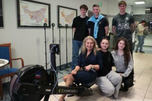 Grupa sześciu osób pozujących przy ergometrze wioślarskim Concept2 w pomieszczeniu przypominającym korytarz lub strefę wspólną. Z przodu, na ergometrze, siedzi blond kobieta w ciemnej sukience; trzyma uchwyt/linę urządzenia i uśmiecha się do aparatu. Obok niej, na podłodze, klęczą dwie młode kobiety: jedna w czarnym stroju z okularami na głowie, druga w szarej bluzie z długimi kręconymi włosami. Z tyłu stoją trzej młodzi mężczyźni: jeden w czarnym T-shircie, drugi w czarnej koszulce z jasnoniebieską bluzą przewieszoną na ramionach, trzeci w ciemnoszarej koszulce z nadrukiem, z widocznymi tatuażami na obu ramionach. Po lewej stronie widać kilka stojaków z tabliczkami z numerami (m.in. 2, 4, 5, 6) oraz dwie duże mapy/wykresy w ramach na ścianie. Po prawej w tle jest część przypominająca bufet lub punkt gastronomiczny, z kilkoma osobami. Na dole po prawej jest podpis: „fot. Magdalena Seredyńska/Uniwersytet Szczeciński”.