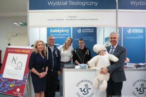 Stoisko promocyjne „Wydział Teologiczny” z logo Uniwersytetu Szczecińskiego oraz grupą pięciu osób pozujących do zdjęcia. W górnej części widać duży napis „Wydział Teologiczny”, a niżej wypunktowane hasła: „teologia” oraz „nauki o rodzinie – doradztwo, coaching, mediacje”. Po lewej stoi czerwony stojak z napisem „PŁYŃ POD PRĄD!” i prostym rysunkiem oraz małym napisem „WYDZIAŁ TEOLOGICZNY”. Na środku jest lada z logo „Uniwersytet Szczeciński” i inicjałami „US”. Na ladzie leżą różne materiały (stos papierów/ulotki), a także mała miska z jasnymi elementami. W tle znajdują się niebieskie ścianki z napisami „Zabłyśnij na Uniwersytecie Szczecińskim” oraz „UNIWERSYTET SZCZECIŃSKI”, a także dodatkowe hasła związane z kierunkami. Po prawej mężczyzna w marynarce trzyma dużą białą pluszową owcę ubraną w białą koszulkę. W prawym dolnym rogu jest podpis: „fot. Magdalena Seredyńska / Uniwersytet Szczeciński”.