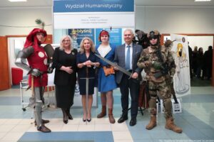 Grupa sześciu osób pozujących do zdjęcia w holu budynku uczelni, na tle dużego baneru z napisem „Wydział Humanistyczny”. W tle widać także inne stoiska i ludzi. Od lewej stoi osoba w stylizowanym stroju rycerskim: metalowa zbroja na rękach i nogach, czerwony kubrak z dużymi białymi kropkami oraz czerwony kaptur/czapka z piórem. Obok stoją dwie kobiety w ciemnych, eleganckich strojach. Dalej stoi młoda osoba w historycznym kostiumie: niebieska tunika, biały kołnierz, czerwone nakrycie głowy i brązowa torba na pasku. Następnie stoi mężczyzna w garniturze; trzyma długi miecz skierowany ukośnie w lewo. Po prawej jest osoba w nowoczesnym umundurowaniu wojskowym w kamuflażu, z hełmem i okularami ochronnymi; trzyma broń. Na banerze pod nagłówkiem „Wydział Humanistyczny” widnieją punkty: „archeologia”, „historia”, „media i cywilizacja”, „stosunki międzynarodowe”. W prawym dolnym rogu zdjęcia jest podpis: „fot. Magdalena Seredyńska/Uniwersytet Szczeciński”.