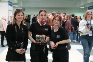 Trzy młode kobiety stojące na pierwszym planie w zatłoczonym holu podczas wydarzenia. Kobieta po lewej ma jasną skórę, brązowe włosy do ramion, na głowie okulary przeciwsłoneczne i czarny strój; na szyi ma pasek aparatu z napisem „Canon”, a w dłoniach trzyma lustrzankę. Kobieta w środku ma jasną skórę, ciemnorude włosy związane z przedziałkiem; ma na twarzy i szyi czerwone plamy wyglądające jak charakteryzacja krwi, czarną koszulkę z białymi literami „SW” i trzyma w rękach przezroczysty pojemnik wypełniony różnymi drobnymi przedmiotami/odpadami. Kobieta po prawej ma jasną skórę, rude włosy, duże okrągłe okulary, czarną koszulkę z białymi literami „SW” i również ma czerwone plamy na dłoniach wyglądające jak charakteryzacja krwi; w rękach trzyma mały ciemny przedmiot. W tle widać wiele osób stojących i rozmawiających w jasnym wnętrzu; po prawej z tyłu stoi kobieta w białej bluzie z identyfikatorem na smyczy i telefonem w dłoni. W lewym dolnym rogu widoczny jest napis: „fot. Maciej Seredyński/Uniwersytet Szczeciński”.