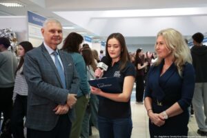 Scena wywiadu podczas wydarzenia uczelnianego, z trzema osobami na pierwszym planie i tłumem w tle. Mężczyzna po lewej ma jasną skórę i siwe włosy, ubrany jest w szarą marynarkę w kratę, białą koszulę i jasnoniebieski krawat, stoi z założonymi rękami i jest zwrócony w stronę aparatu. Kobieta w środku ma jasną skórę i proste, ciemnobrązowe włosy do ramion; trzyma mikrofon oraz granatową teczkę, ma na sobie czarną koszulkę polo z białym logo „UNIWIZJA” na piersi, wygląda jak prowadząca wywiad. Kobieta po prawej ma jasną skórę i długie, falowane blond włosy; ubrana jest w granatową sukienkę lub kostium z paskiem, ma zegarek i bransoletkę, stoi bokiem i jest zwrócona w stronę prowadzącej. W tle widać wiele osób oraz stoiska z tablicami; na jednej tablicy częściowo widoczny napis zaczynający się od „Wydział Hu…”. W prawym dolnym rogu jest podpis: „fot. Magdalena Seredyńska/Uniwersytet Szczeciński”.