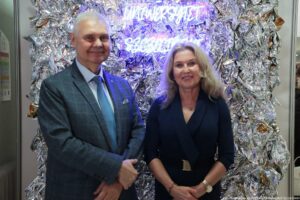 Dwie osoby pozujące do zdjęcia na tle srebrnej, pogniecionej, błyszczącej folii z neonowym napisem. Po lewej stronie stoi starszy mężczyzna o siwych włosach, w szarym garniturze, jasnej koszuli i niebieskim krawacie; ma lekki uśmiech i ręce złożone z przodu. Po prawej stoi kobieta o jasnej cerze i długich, blond włosach, w ciemnej, eleganckiej sukience z długimi rękawami; ma delikatny uśmiech, naszyjnik, bransoletkę i zegarek, a ręce trzyma złożone z przodu. W tle u góry widoczny jest świecący na fioletowo napis „UNIWERSYTET”, a poniżej rozmyte „SZCZECIŃSKI”. W prawym dolnym rogu znajduje się podpis: „fot. Magdalena Seredyńska / Uniwersytet Szczeciński”.