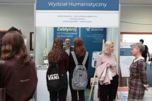 Stoisko informacyjne z dużym napisem „Wydział Humanistyczny”. Pod spodem, mniejszym drukiem: „dziennikarstwo i komunikacja społeczna”, „filologia polska”, „studia pisarskie”, „italianistyka”. Na pierwszym planie stoją osoby rozmawiające przy stoisku. W centrum dwie młode kobiety odwrócone tyłem, z plecakami (jeden czarny z napisem „VANS”, drugi w czarno-białą kratkę). Przed nimi mężczyzna w średnim wieku rozmawia i uśmiecha się, stojąc za ladą stoiska. Po prawej dwie kobiety rozmawiają ze sobą: jedna blondynka w jasnoróżowej bluzce trzyma w ręku jasną kurtkę, druga ma krótkie siwe włosy i sukienkę w kratę. W tle widoczne są granatowe plansze z napisem „Zabłyśnij na Uniwersytecie Szczecińskim” oraz „Uniwersytet Szczeciński”. Po prawej stronie widać też fragment innego stoiska z napisem zaczynającym się od „Wydział Ekono… Finansów… i Zarządz…”. W prawym dolnym rogu jest podpis fotograficzny: „fot. Magdalena Seredyńska/Uniwersytet Szczeciński”.