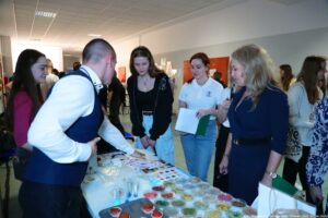 Grupa osób stojących przy stole z materiałami do doświadczeń w jasnym korytarzu. Na pierwszym planie stół jest przykryty białym obrusem i zastawiony wieloma przezroczystymi, okrągłymi pojemnikami (wyglądają jak płytki Petriego) z kolorowymi substancjami: czerwonymi, żółtymi, zielonymi i brązowymi. Widać też kilka plastikowych probówek i drobny sprzęt laboratoryjny. Przy stole stoją głównie młode osoby: z lewej dziewczyna z długimi, ciemnymi włosami w bordowej bluzce; obok mężczyzna w białej koszuli i ciemnej kamizelce pochyla się nad stołem; w środku dziewczyna z brązowymi włosami w czarnej bluzie i jasnych spodniach patrzy na eksponaty; obok kobieta w białej koszulce polo trzyma kartki i zieloną teczkę. Po prawej stoi blondynka w granatowej sukience z paskiem, również z zieloną teczką. W tle jest więcej osób i jasny sufit z panelami oświetleniowymi. W prawym dolnym rogu znajduje się podpis: „fot. Magdalena Seredyńska/Uniwersytet Szczeciński”.