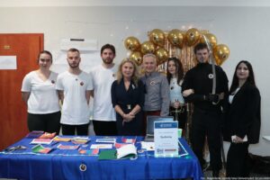 Grupa ośmiu osób stojących za stołem promocyjnym. Na zdjęciu są cztery kobiety i czterech mężczyzn, ustawieni w jednym rzędzie, pozują do wspólnej fotografii. Kilka osób ma na koszulkach lub ubraniach okrągłe przypinki z podobnym symbolem. Jedna osoba po prawej stronie trzyma długą, zakrzywioną szablę w pochwie lub z osłoną rękojeści. Przed nimi stoi stół przykryty niebieskim obrusem. Na blacie leżą małe flagi różnych państw, ulotki i materiały papierowe, długopisy oraz otwarty notes. Jest też laptop oraz stojąca tabliczka z napisem „Uniwersytet Szczeciński”, „Koło Naukowe Sydonia”, „Wydział Nauk Społecznych”, „Dni otwarte”. W tle po prawej stronie wisi złota kurtyna z frędzli, a wokół niej jest wiele złotych balonów. Po lewej stronie widać czerwone drzwi z kartką informacyjną. W prawym dolnym rogu jest podpis: „fot. Magdalena Seredyńska/Uniwersytet Szczeciński”.