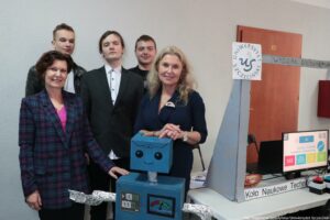 Grupa pięciu osób stojących przy stole z niebieskim modelem robota z kartonu. Na pierwszym planie są dwie kobiety: po lewej w kraciastej marynarce w odcieniach fioletu z różową bluzką, po prawej w granatowej sukience, z długimi jasnymi włosami. Za nimi stoją trzej młodzi mężczyźni w ciemnych ubraniach; jeden ma na sobie marynarkę i białą koszulę. Na stole stoi kartonowy robot: niebieska, kwadratowa głowa z narysowanymi dużymi oczami i uśmiechem, niebieski tułów oraz „ramiona” z folii aluminiowej wystające na boki. Obok, po prawej stronie stołu, jest duży szary kartonowy element przypominający pylon lub znak. Na nim widać napis „UNIWERSYTET SZCZECIŃSKI” oraz na poziomej tabliczce „WYDZIAŁ EKONOMII, FINANSÓW I ZARZĄDZANIA”. Na przedniej krawędzi stołu jest napis „Koło Naukowe Techn…”, ucięty na końcu kadru. Po prawej na stole stoi monitor komputera z kolorowym slajdem; w prawym górnym rogu ekranu jest „0/3”, a na slajdzie widać „WYDZIAŁ EKONOMII, FINANSÓW I ZARZĄDZANIA”. W prawym dolnym rogu zdjęcia jest podpis: „fot. Magdalena Seredyńska/Uniwersytet Szczeciński”.