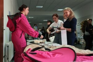 Scena z wydarzenia w pomieszczeniu, z kilkoma osobami stojącymi przy stole. W lewym planie stoi młoda kobieta o jasnej skórze w intensywnie różowym kombinezonie, z czarnymi elementami na plecach, pochylona nad stołem. Na stole leżą różne drobne przedmioty i materiały (m.in. papier, pojemniki i ulotki), a na pierwszym planie znajduje się rozłożony różowy przedmiot z czarną lamówką (wygląda jak torba lub pokrowiec). Po prawej stronie dwie kobiety o jasnej skórze trzymają teczkę i kartki; jedna ma blond włosy i granatową sukienkę, druga ma brązowe włosy i białą koszulkę polo. W tle widać więcej ludzi, a przy ścianie są złote balony i drobne światełka dekoracyjne. W prawym dolnym rogu jest podpis: „fot. Magdalena Seredyńska/Uniwersytet Szczeciński”.