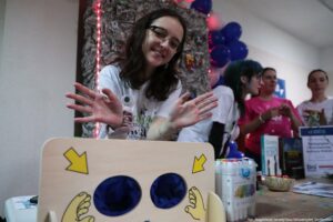 Stanowisko na wydarzeniu w pomieszczeniu, z młodą kobietą na pierwszym planie uśmiechającą się do aparatu i trzymającą dłonie uniesione przed sobą. Kobieta ma jasną skórę, długie ciemne włosy i okulary. Ma na sobie białą koszulkę z nadrukiem i przypiętą plakietką/imiennikiem. Na stole stoi drewniana skrzynka/urządzenie z dwiema okrągłymi dziurami wyłożonymi granatowym materiałem; na frontowej ściance są rysunki dłoni oraz żółte strzałki wskazujące otwory. Obok leżą/zostały postawione pojemniki z mazakami i różne materiały informacyjne. W tle widać kilka osób przy stoisku (również w koszulkach), a nad nimi dekoracje z balonów w odcieniach niebieskiego i różowego oraz światełka. Na zdjęciu jest też drobny podpis w prawym dolnym rogu: „fot. Magdalena Seredyńska/Uniwersytet Szczeciński”.