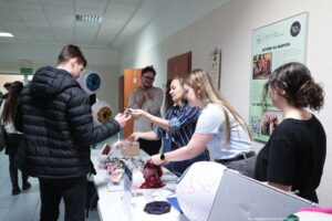 Korytarz w budynku, przy ścianie stoi stolik z materiałami edukacyjnymi i modelami anatomicznymi oczu. Kilka osób rozmawia przy stoliku; młody mężczyzna w czarnej puchowej kurtce stoi po lewej i odbiera coś (prawdopodobnie ulotkę lub mały przedmiot) od kobiety stojącej za stolikiem. Na stole leżą drobne przedmioty i breloczki, miska z zawartością, czerwony woreczek, a także duży biały model gałki ocznej z czerwonymi naczyniami oraz modele przekrojów oka. W tle na ścianie wisi tablica z numerem „4” oraz duży plakat z nagłówkiem „JESTEŚMY DLA MŁODYCH!” i logo „YOUNG POWER”; na końcu korytarza jest zielony znak „WYJŚCIE”. W prawym dolnym rogu widoczny jest napis: „fot. Magdalena Seredyńska/Uniwersytet Szczeciński”.