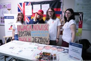 Stoisko promocyjne na uczelni z trzema osobami stojącymi za stołem oraz dużą tablicą z ręcznie napisanym hasłem. Na środku trzymany jest kartonowy plakat z kolorowymi literami: „UWAGA! NOWOŚĆ ZOSTAŃ NAUCZYCIELEM TŁUMACZEM DWÓCH JĘZYKÓW”. Za osobami wisi dekoracja złożona z flag (widać motywy Wielkiej Brytanii, Niemiec oraz Francji) i kilka wydrukowanych kartek z informacjami. Na stole leżą ulotki i plansze informacyjne. Po prawej stronie stoi tabliczka z logo „Uniwersytet Szczeciński” oraz tekstem: „Koło Naukowe Młodych Germanistów”, „Wydział Humanistyczny”, „Dni otwarte”. Na stole jest też metalowa puszka z motywem flagi Wielkiej Brytanii, słodycze oraz kilka papierowych twarzy na patyczkach jako rekwizyty. W lewym dolnym rogu widoczny jest podpis: „fot. Magdalena Seredyńska/Uniwersytet Szczeciński”.